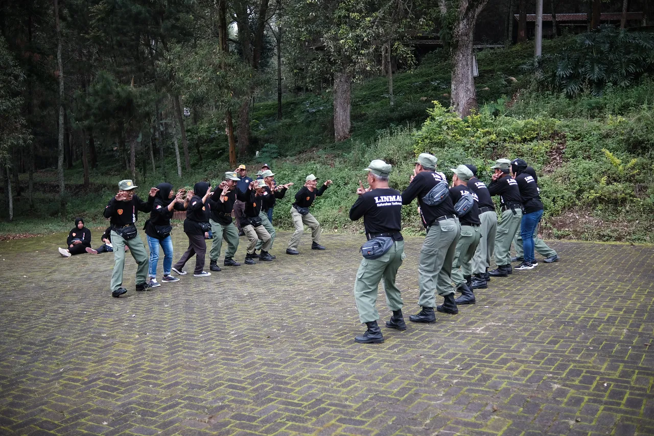 Rombongan peserta beragam usia mengikuti permainan di pelataran paving berbatasan langsung dengan hutan pinus
