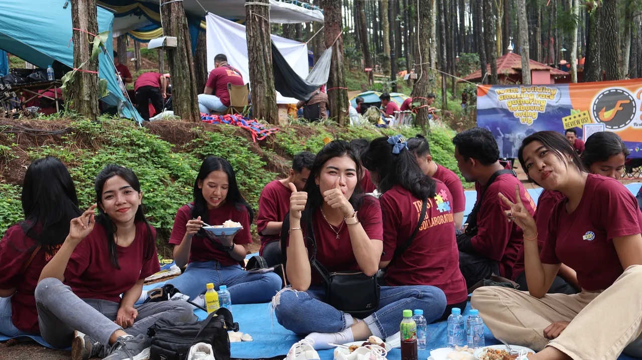 Peserta makan siang beralaskan terpal di area hutan pinus dengan tenda dan terpal peneduh terpasang di belakang