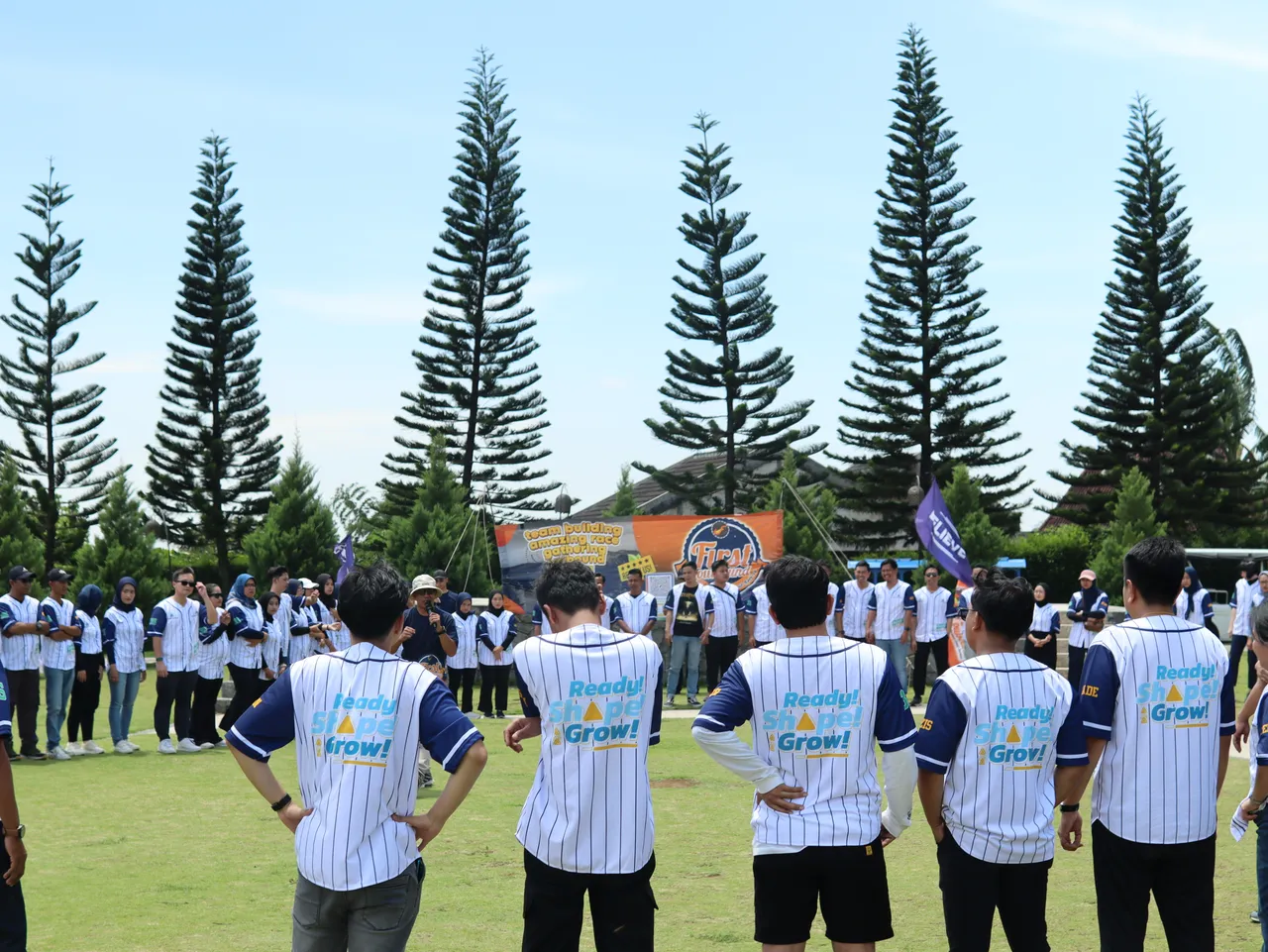 Fasilitator memberi pengarahan kepada peserta yang berdiri melingkar di lapangan rumput dengan atap bangunan terlihat di kejauhan